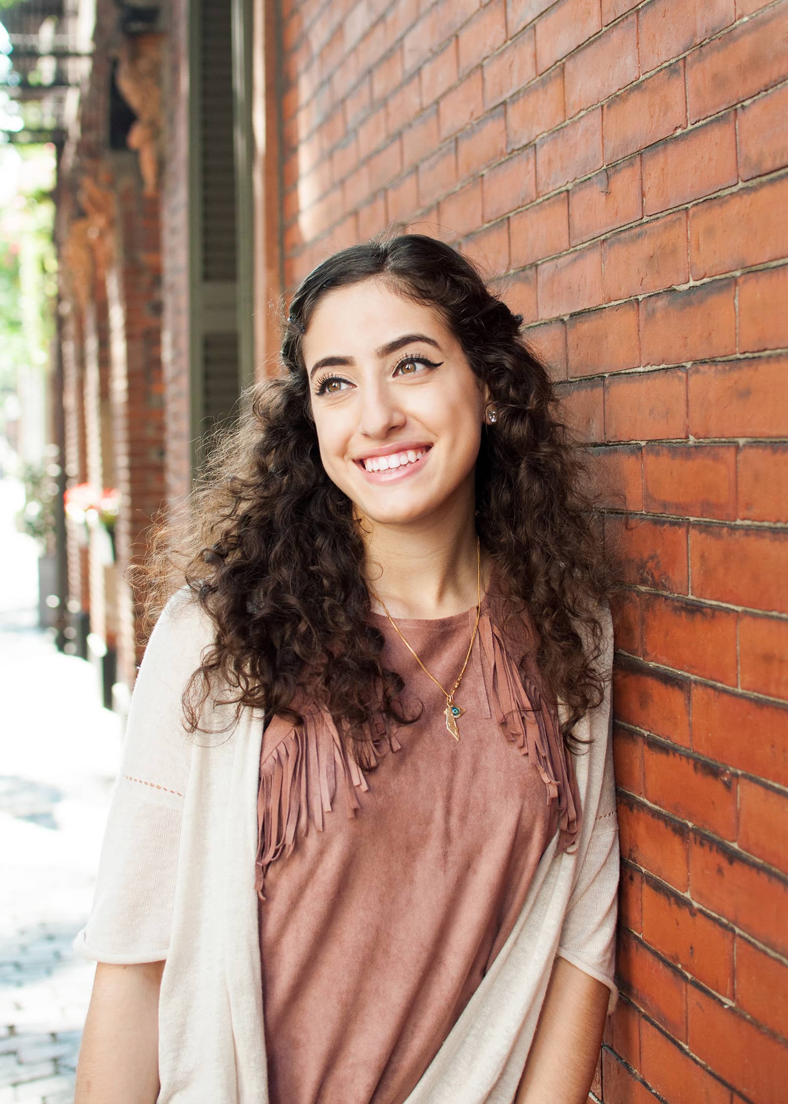 Natural-light portrait of a smiling woman with long curly hair posing beside a red brick wall in an urban setting.